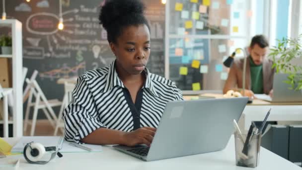 Retrato de una joven afroamericana usando un portátil y escribiendo datos en un cuaderno en la oficina — Vídeos de Stock