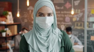 Tıbbi maske takan Müslüman bir ofis çalışanının yavaş çekim portresi iş yerinde kameraya bakıyor.