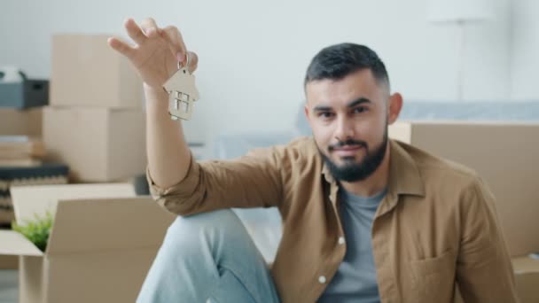 Портрет красивого ближневосточного мужчины с ключом, сидящего на полу с картонными коробками на заднем плане — стоковое видео
