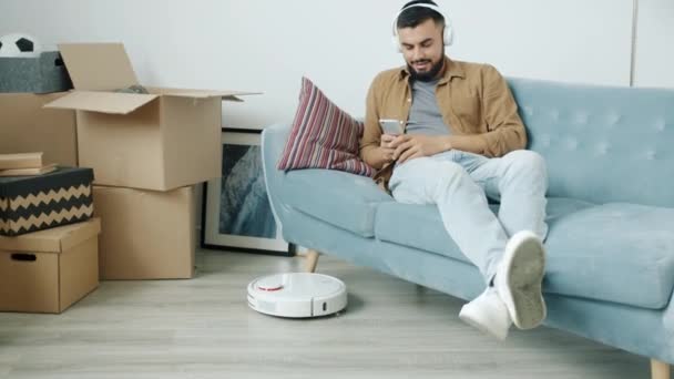Joyful misto raça homem se divertindo com smartphone enquanto robótico aspirador de pó de pó chão na nova casa — Vídeo de Stock