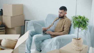 Orta Doğulu genç bir adamın portresi. Dizüstü bilgisayar kullanıyor. Kanepede karton kutularla oturuyor.