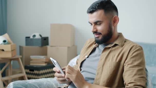 Hombre alegre de Oriente Medio usando la pantalla táctil del teléfono inteligente sentado en un nuevo apartamento con cajas de cartón — Vídeos de Stock