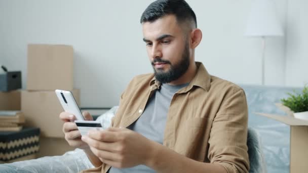 Młody Arab człowiek zakupy online palyment ze smartfonem w nowym mieszkaniu z kartonowymi pudełkami — Wideo stockowe