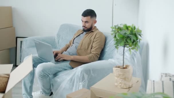 Arab guy freelancer working with laptop sitting on couch in apartment full of cardboard boxes — Stock Video