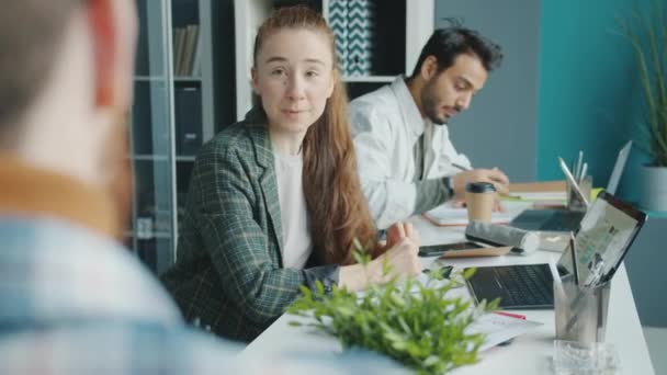 Jonge zakenvrouw in gesprek met collega 's die documenten aannemen die in een gedeeld kantoor aan een bureau werken — Stockvideo