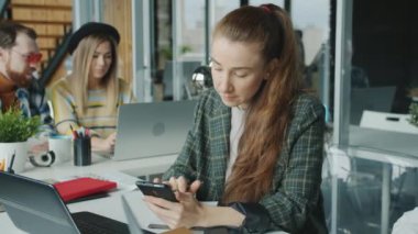 Bilgisayarla çalışan ve kapalı bir ofiste akıllı telefon kullanan genç bir kadın.