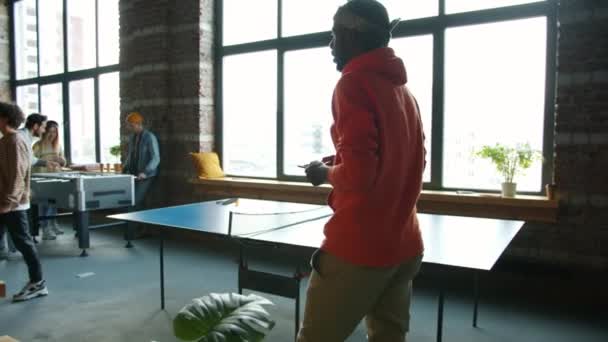 Молодой афро-американский бизнесмен, входящий в творческий офис, наблюдает за коллегами, работающими и играющими в настольный футбол — стоковое видео