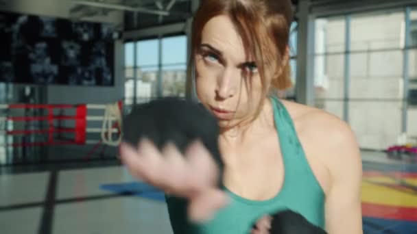 Kobieta zawodniczka z owiniętymi rękami porusza ramionami rzucając ciosy i patrząc na kamerę w siłowni — Wideo stockowe