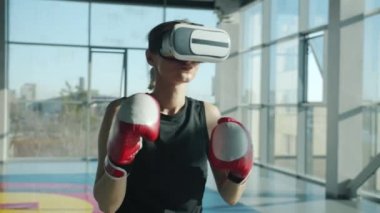 Modern spor salonunda sanal gerçeklik gözlüğü ve boks eldiveni takan genç bir kadın.