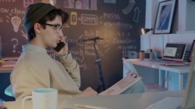 Erkek çalışan cep telefonuyla konuşuyor ve karanlık ofiste çalışan kağıtları kontrol ediyor.