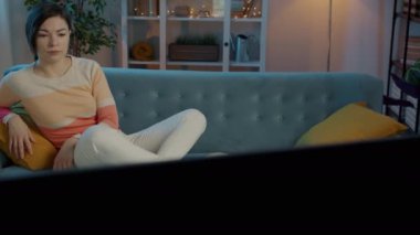 Yorgun ve uykulu genç bir kadın kanepede dinleniyor ve akşamları evde televizyon izliyor.