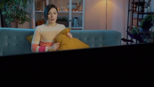 Verängstigte junge Frau vor dem Fernseher runzelt die Stirn und versteckt sich hinter Kopfkissen, wenn sie nachts auf dem Sofa sitzt — Stockvideo