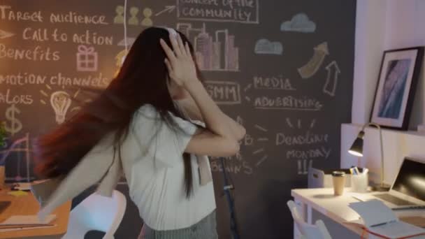 Hermosa chica asiática empleada bailando en el lugar de trabajo con auriculares disfrutando de la música por la noche — Vídeos de Stock