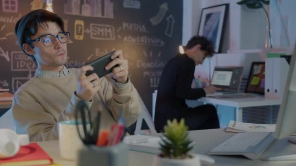 Glädjande anställd njuter av videospel spela på jobbet medan kollega arbetar i bakgrunden — Stockvideo