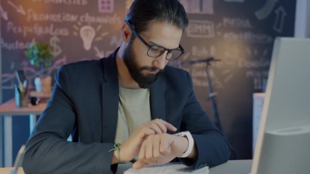 Middle Eastern entrepreneur using smart watch swiping screen sitting at desk in dark office — Stock Video