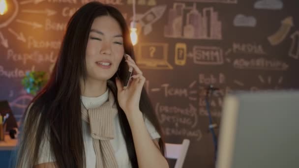 Feliz mujer de negocios asiática charlando en el teléfono móvil trabajando en interiores en la oficina creativa por la noche — Vídeos de Stock