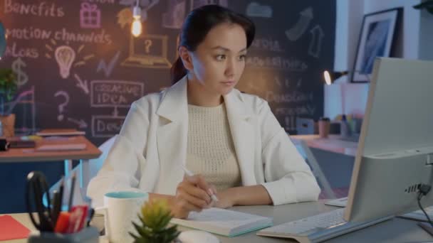 Mulher de negócios asiática bonita tomar notas olhando para monitor de computador trabalhando à noite no escritório criativo — Vídeo de Stock