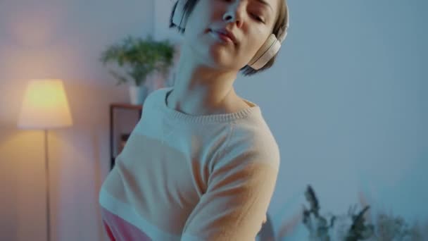 Πορτρέτο της χαριτωμένης φοιτήτρια χορό στο σκοτεινό διαμέρισμα φορώντας ακουστικά — Αρχείο Βίντεο