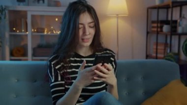 Mutlu öğrenci gülüyor ve akıllı telefon kullanıyor. Geceleri kanepede oturuyor.
