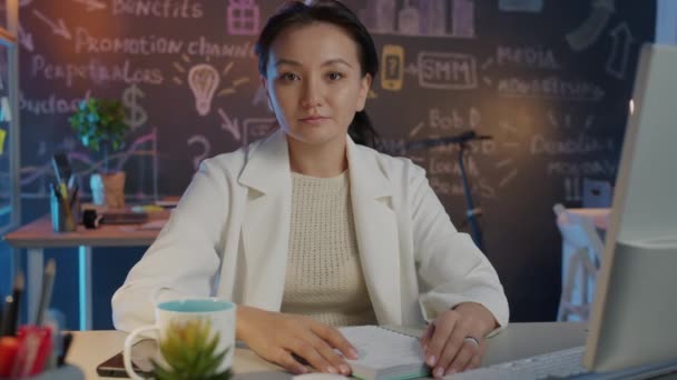 Asyalı ciddi bir iş kadınının karanlık yaratıcı ofisinde masa başında kameraya bakması ağır çekimde. — Stok video