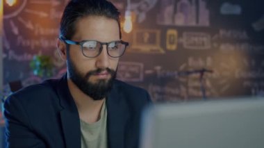 Karanlık ofiste bilgisayar kullanan çekici Orta Doğulu adam online işlere odaklanmış.