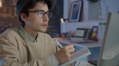 Geceleri ofiste çalışan bilgisayar monitörüne bakan düşünceli bir çalışan notlar yazıyor.
