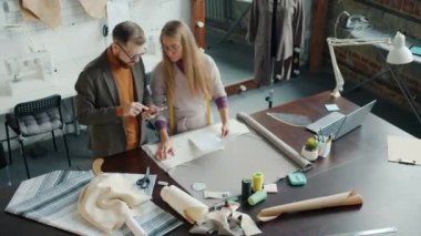 Erkek ve kadın terziler elbise çizimlerine bakıyor ve stüdyoda birlikte çalışırken akıllı telefon kullanıyorlar.
