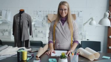 Modern stüdyoda çalışma masasında kameraya bakıp gülümseyen başarılı bir terzi.