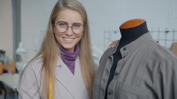 Portret van een mooie jonge vrouw in de buurt van een pop met handgemaakt kledingstuk dat binnen glimlacht in de werkplaats — Stockvideo