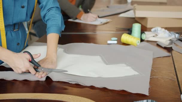 Close-up van kleermakers handen snijden stof maken van nieuwe kleding terwijl collega werkt op de achtergrond — Stockvideo