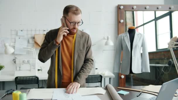 Kreatywny krawcowa facet rozmawia przez telefon komórkowy i patrząc na szkice pracy w studio sam — Wideo stockowe
