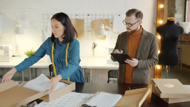 Jonge vrouw verpakking van nieuwe kleren in kartonnen dozen, terwijl de eigenaar van het bedrijf met behulp van tablet — Stockvideo