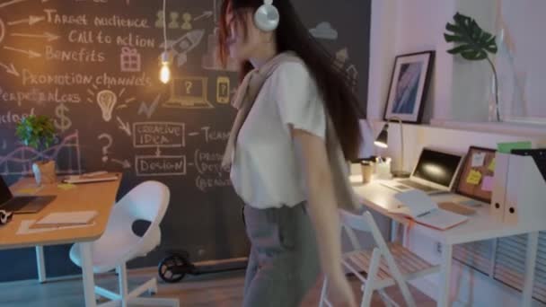 Carefree Asian businesswoman wearing headphones dancing in creative office at night — 图库视频影像