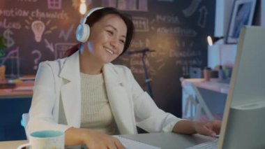 Güzel Asyalı kadın kulaklıkla müzik dinliyor ve geceleri yaratıcı ofiste bilgisayarla çalışıyor.