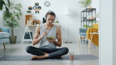Evde yoga minderinde oturan, kulaklık takan, salata yiyen yakışıklı adamın yavaş çekimde portresi.