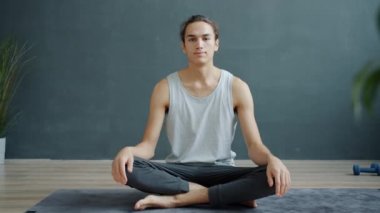 Erkek yoga eğitmeninin yavaş çekim portresi güzel bir stüdyoda mindere oturup kameraya bakıyor.
