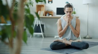 Modern apartmandaki yoga minderinin üzerinde oturup namaste 'de ellerini hareket ettirerek meditasyon yapan melez bir adamın yavaş hareketi.
