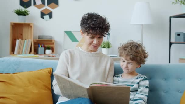 Молодая мать читает интересную книгу милому мальчику, воспитывающему ребенка дома — стоковое видео