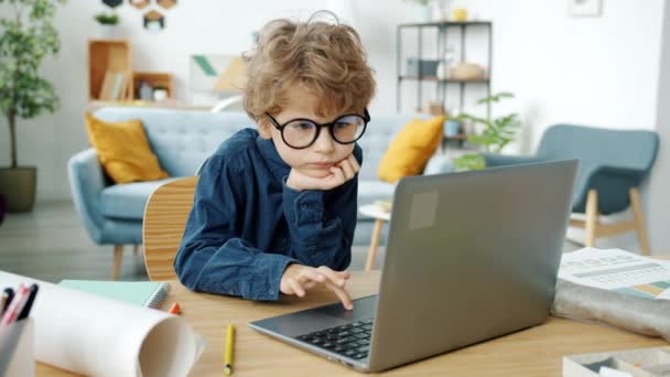 Έξυπνο μικρό αγόρι φοράει γυαλιά χρησιμοποιώντας φορητό υπολογιστή στο τραπέζι στο διαμέρισμα — Αρχείο Βίντεο