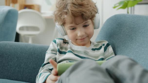 Cámara lenta del niño tocando la pantalla del teléfono inteligente y escuchando audiolibros a través de auriculares en casa — Vídeo de stock
