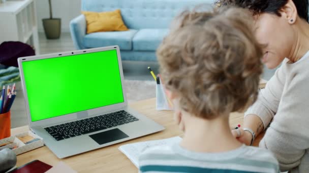 Cuidado de la madre ayudando a su hijo con la escritura de tareas mirando a la pantalla verde croma clave portátil en casa — Vídeos de Stock