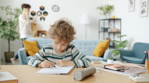 Menino escrevendo estudando fazendo lição de casa enquanto a mãe falando no celular em casa — Vídeo de Stock