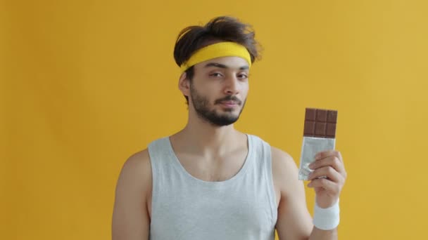 Портрет серьезного арабского спортсмена, нюхающего шоколад, а затем отказывающегося от сахара на желтом фоне — стоковое видео