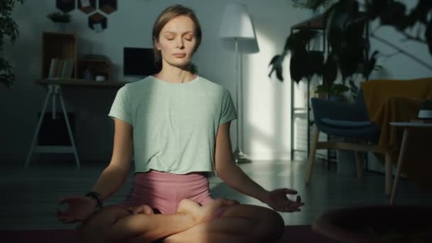 Schöne junge Frau meditiert allein zu Hause und sitzt in Lotusposition auf einer Yogamatte und genießt Entspannung — Stockvideo