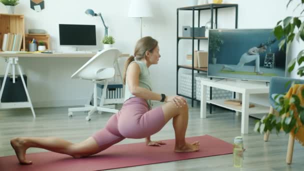 Jovem senhora em cima e shorts aprendendo ioga asanas assistindo tutorial na TV exercitando em casa no tapete — Vídeo de Stock