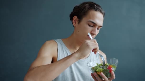 Портрет счастливого молодого спортсмена, поедающего полезный овощной салат на сером фоне — стоковое видео