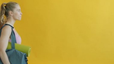 Yoga minderli ve su şişeli spor çantası taşıyan genç bir kadının yavaşça yürümesi.
