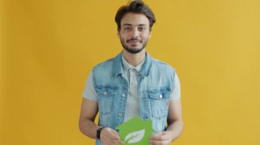 Gülümseyen Orta Doğulu adam elinde yeşil enerji sembolü eviyle sarı arka planda yaprak tutuyor.