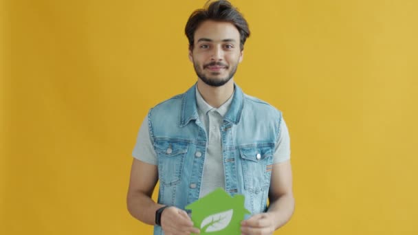 Sonriente hombre de Oriente Medio sosteniendo la casa símbolo de energía verde con hoja sobre fondo amarillo — Vídeos de Stock