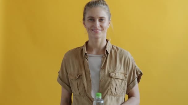 Retrato de una joven bebiendo agua de una botella de vidrio reutilizable sobre fondo amarillo — Vídeos de Stock